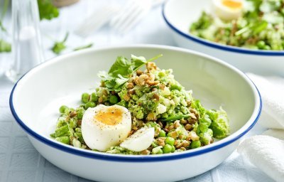 Een wit emaille bordje met blauwe rand is gevuld met de linzensalade. Het is een salade rijk aan groene groenten. Het gerecht is afgewerkt met een gekookt eitje. 