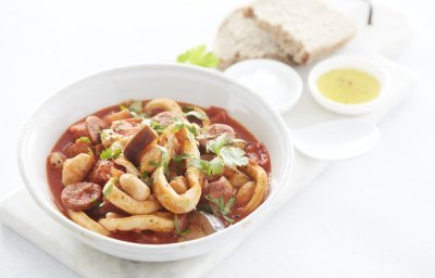 Een goed gevuld wit bord met het stoofpotje staat klaar om aan te vallen. De kleur is rood van de vele tomaten en chorizo die zijn gebruikt. Het bordje staat op een marmere snijplank waar ook brood en een klein potje olijfolie op staat. 