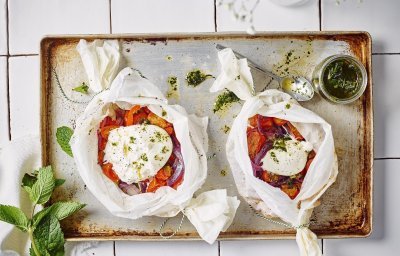 Twee papillotten liggen op een ovenplaat. De papillot is gemaakt in een wit boterpapiertje waarin de groenten een mooi contrast in geven. Denk maar aan de rode paprika's en tomaten. De burrata ligt er bovenop en de muntolie staat in een bokaaltje naast de papillotten.