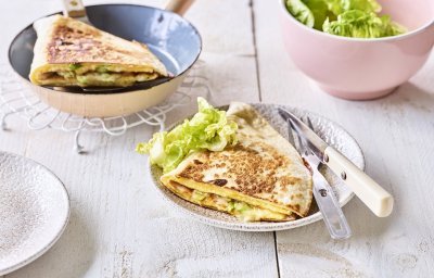 Een krokante tortilla ligt in een bordje klaar om aan te vallen. Hij is gevuld met een omelet, passendale kaas, gandaham en lente-ui. De tweede quesadilla ligt nog in de pan en staat ook op de tafel. 