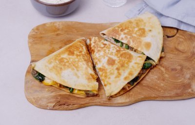 Een klaargemaakte quesadilla ligt op een houten snijplank. De quesadilla is in 3 gesneden, hij is gevuld met vlees, spinazie, zwarte bonen en kruiden. 