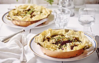 Aan tafel! Quiche van filodeeg met geitenkaas en rode biet staan klaar op tafel. De quiches zitten in een kopperen schaaltje en staan op een wit bord. Het bestek en de witte servette liggen langs het bord. Op tafel staan ook waterglazen en een glazen peper- & zoutvat.