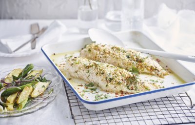 De gegrilde roodbaars uit de oven ligt op een witte bakplaat en staat op een rooster. De crumble zorgt voor een zomerse sfeer in het gerecht. Naast de ovenschaal staat een bordje met wat aardappelsalade uitgeschept. 