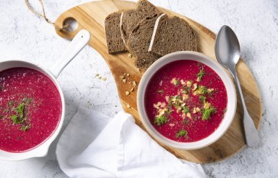 Soep van rode biet kleurt natuurlijk rood en smaakt naar nog! Ze is geserveerd in twee witte kommetjes en staat op een houten snijplank met enkele bruine sneden brood bij. 