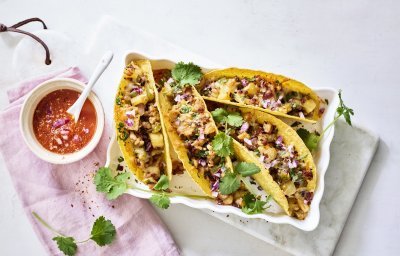 Vier tacos liggen klaar in een ovenschaal om aan te vallen. De pittige tomatendip staat links van de schotel op een roze servette. Het gerechtje is afgewerkt met verse korianderblaadjes.