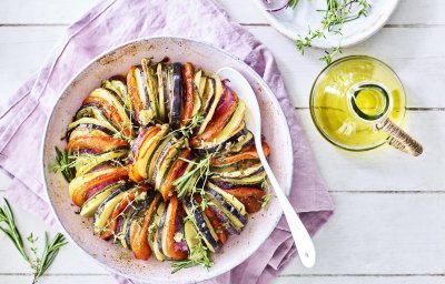 Prachtige tian boordevol met schijfjes van zomerse groenten en aardappel. Hij staat op een roze servette en een side bordje met rode ui en verse kruiden. Zomer in een foto!