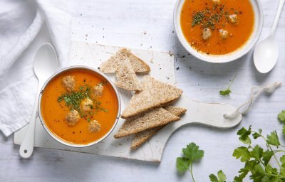 Heerlijk gemaakte tomatensoep staat uitgeschenkt op tafel in twee soepborden. De balletjes drijven in de soep en is afgewerkt met peterselie. Brood en lepels liggen langs het bord, klaar om te slurpen.