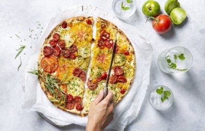 Een kleurrijke tomatentaart staat op tafel op een wit boterpapiertje. Ze wordt aangesneden door een onbekende vrouw met een koksmes. Rechtsboven liggen nog enkele tomaten om ze extra in de picture te zetten. 