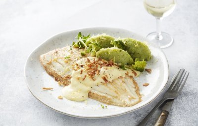 Heerlijk gebakken tongschar ligt op een wit bord met de broccoliepuree naast. Het sausje is over de vis gedresseerd met enkele gefrituurde uitjes. Bestek en een glaasje witte wijn werken het beeld af. 