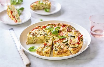 Een heerlijke aardappeltortilla ligt aangesneden op een groot wit bord. Er werden al twee stukjes uitgesneden en liggen geserveerd op kleine bordjes en afgewerkt met basilicum. Er staan licht gekleurd roze glazen langs het gerecht om de foto nog meer kleur te geven.