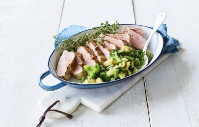 Een heerlijk varkenshaasje uit de oven ligt in een blauwe ovenschaal, vergezeld met groene groenten en verse tijm. 