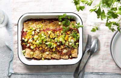 Enchiladas liggen mooi op een rijtje in een witte ovenschotel. Hierop werd tomatensaus geserveerd en gemalen kaas, in de oven. Versgesneden lente-uitjes werden als afwerking toegevoegd aan het gerecht. 