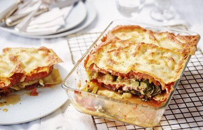 De lasagne staat op tafel in een glazen doorzichtige ovenschotel. Hij is aangesneden zodat je mooi de laagjes ziet van pasta, groenten en saus. De portie die is uitgeschept ligt op een bordje er naast.