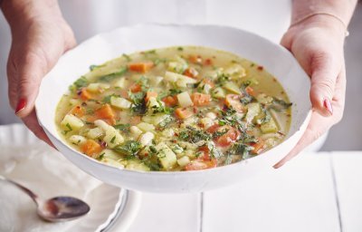 Een vegetarische minestrone met venkel, witte bonen, aardappel en verse kruiden geserveerd in een diep wit soepbord. Een vrouw met mooi rood gelakte nagels draagt het bord. 