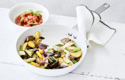 Vegetarische wok van aubergine en courgette staat klaar om aan te vallen in een witte wokpan. Er is een potje gevuld met tomatenblokjes, basilicum, azijn en olijfolie waarmee je je gerecht een extra frisse toets geeft.