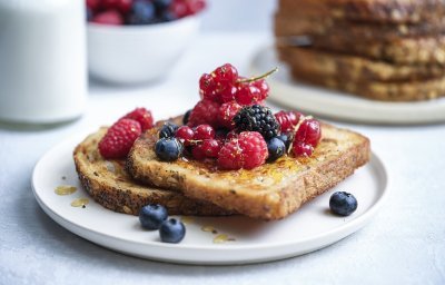 Twee wentelteefjes liggen op elkaar op een wit bord. Ze zijn mooi krokant gebakken en werden geserveerd met zomerfruit, in dit geval aardbeien, bosbessen, frambozen en rode bessen. In de achtergrond zie je de andere stapel wentelteefjes liggen, een kan melk en een bordje met nog extra zomerfruit.