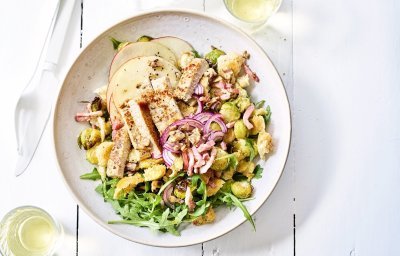 Winterse panzanella met ovengeroosterde spruitjes, versgesneden appel en gegrilde kalfsoester in een diep wit bord. Wit bestek en wijn staat langs het bord, klaar om aan te vallen.