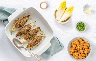 Gevulde kroppen witloof met kruidig gehakt liggen in een ovenschotel. De schotel staat op een lichtblauwe doek met kruidige aardappelen in kommetje langs. 