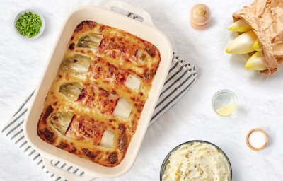 Gegratineerde witloofrolletjes opgerold in gekookte ham liggen mooi naast elkaar in een ovenschotel. De kaassaus is laat de rolletjes net bovenkomen. Er staat een pot heerlijke puree klaar om aan te vallen. 