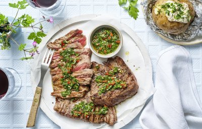 Het heerlijk stukje vlees ligt op een groot wit rond bord. Het al voor de helf in reepjes gesneden en bedruppeld met de Aziatische chimichurri. Er ligt een gepofte aardappel langs en twee glaasjes rode wijn. 