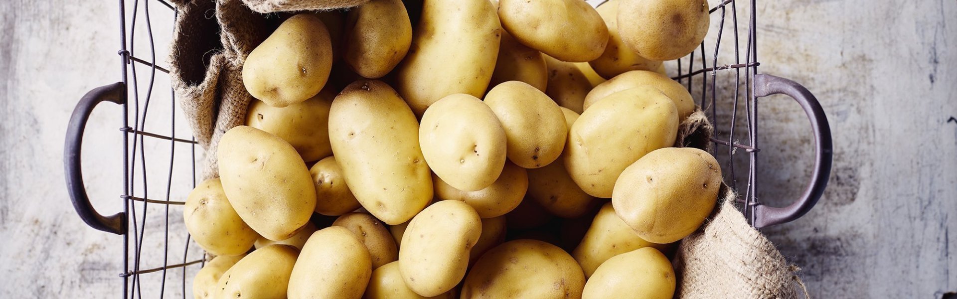 Aardappelen liggen in ijzeren mand met bruine linnen doek onder. De mand staat op een grijze gietbeton.