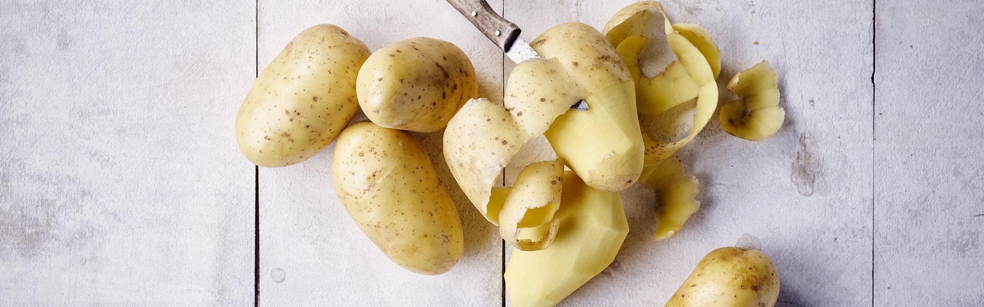 Aardappelen liggen op oude houten tafel. Er zijn al enkele geschild en eentje half waarbij het aardappelmesje er bij ligt.