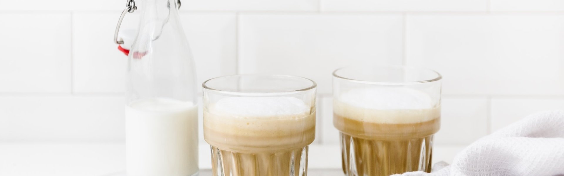 Latte geschonken in doorzichtige glazen. Er staat een klein halfvol melkflesje naast. Alles is gepresenteerd op een wit dienblad met koperen lepeltje, linnen theedoek en enkele koffiebonen ter versiering. 