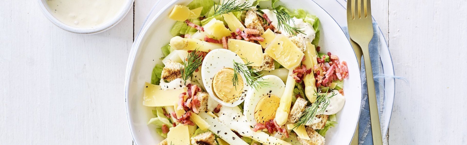 Een rijkgevulde salade staat klaar op tafel. Het witte bord is gevuld met sla, gekookte eitjes, stukjes asperges en kaasschilfers. Dat is de lente proeven in je bord, als je het mij vraagt!