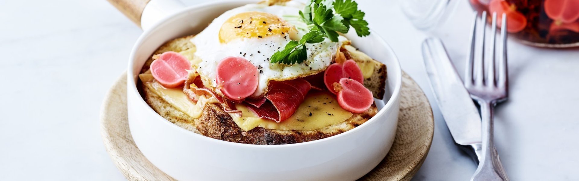 Croute au fromage met Averbode kaas in een witte pan, spiegeleitje er bovenop gepresenteerd met een takje verse peterselie. Op de achtergrond staan gepekelde radijzen in glazen pot.