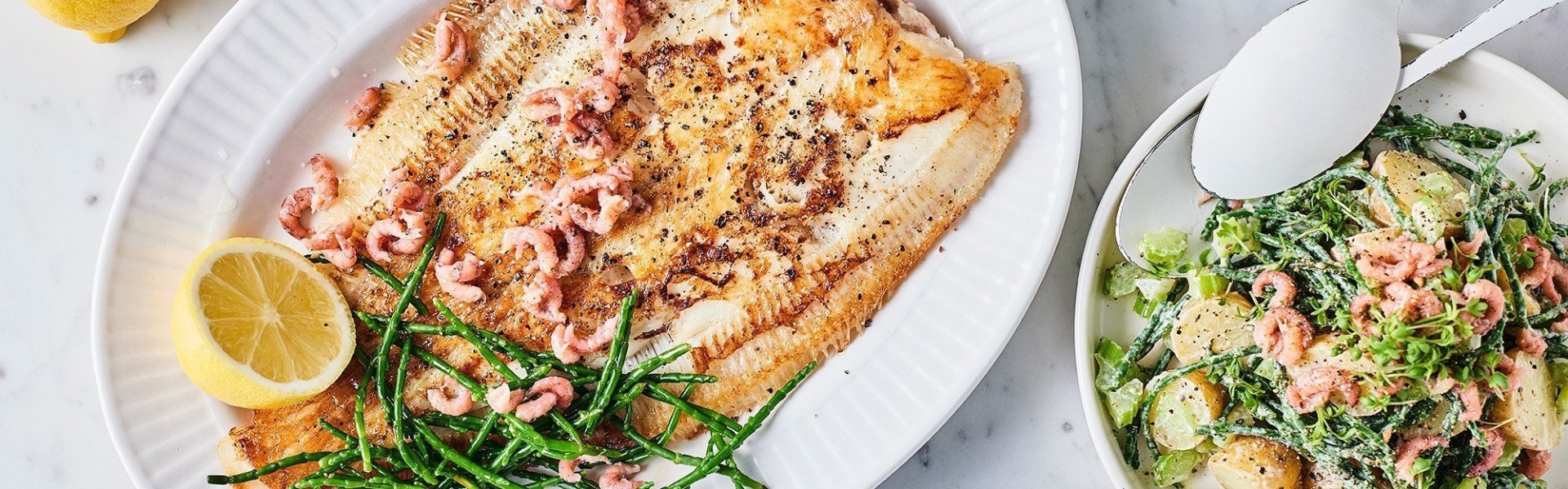 Een heerlijk krokant visje afgewerkt met zeekraal ligt op een groot bord. Er zijn enkele garnaaltjes aan toegevoegd die mooi roze blinken. De aardappelsalade staat er langs in een witte kom met witte lepel. Om af te werken ligt er 1 citroen doormidden gesneden.