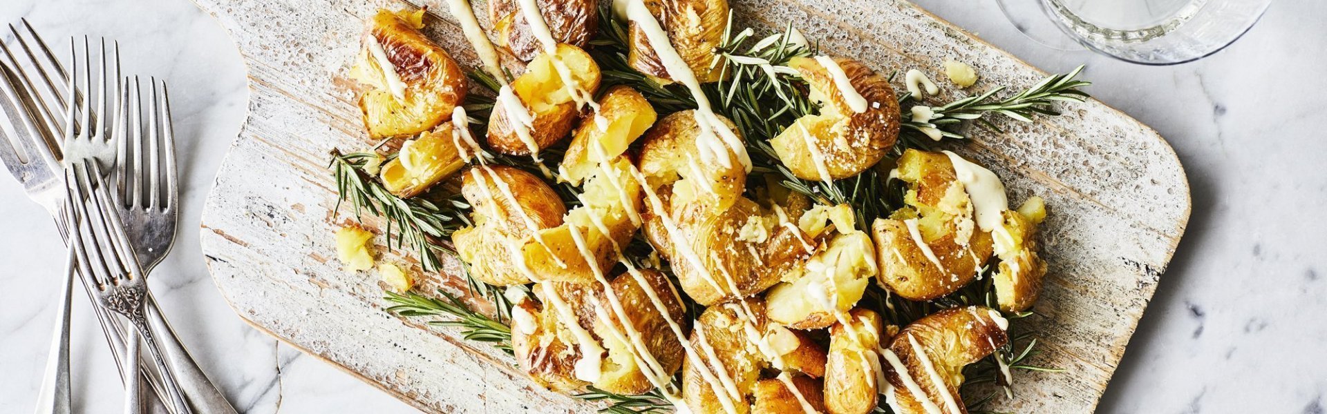 Gecrushte aardappelen met picklessausje Eens een patatje in een feestjas op tafel? Serveer deze ovenaardappeltjes en ontvang veel "mmmmm-lekker complimentjes". Deze gecrushte aardappeltjes zijn geserveerd op een houten plank met een heerlijk picklessausje.