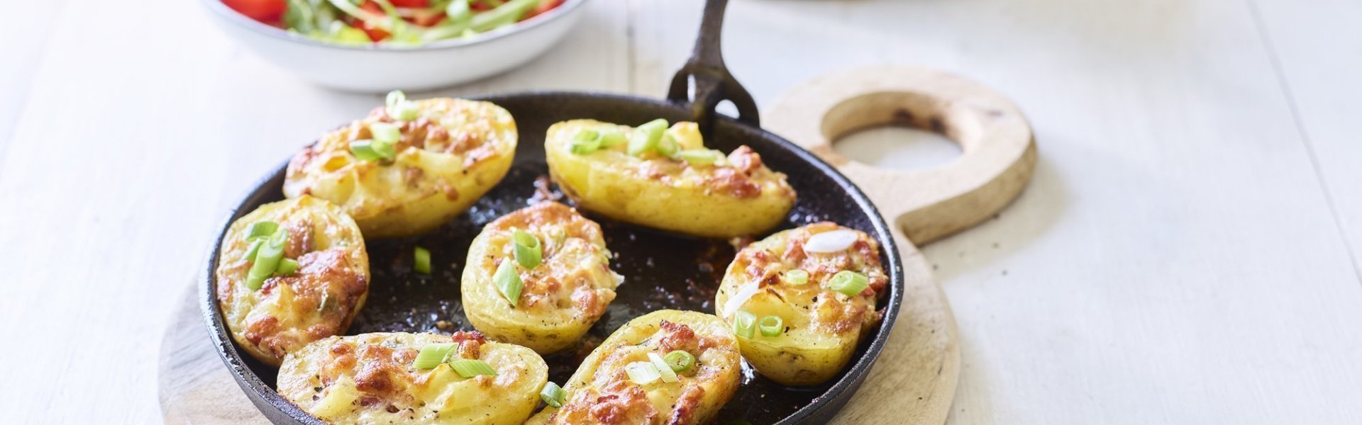 Gevulde aardappelen met spek en Flandrien kaas op de barbecue 7 gevulde aardappelen liggen op een zwarte vuurvaste pan, ze zijn gevuld met aardappelkruim, kaas en spekblokjes. Er ligt een handdoek achteraan in beeld en een slaatje met tomaatjes.