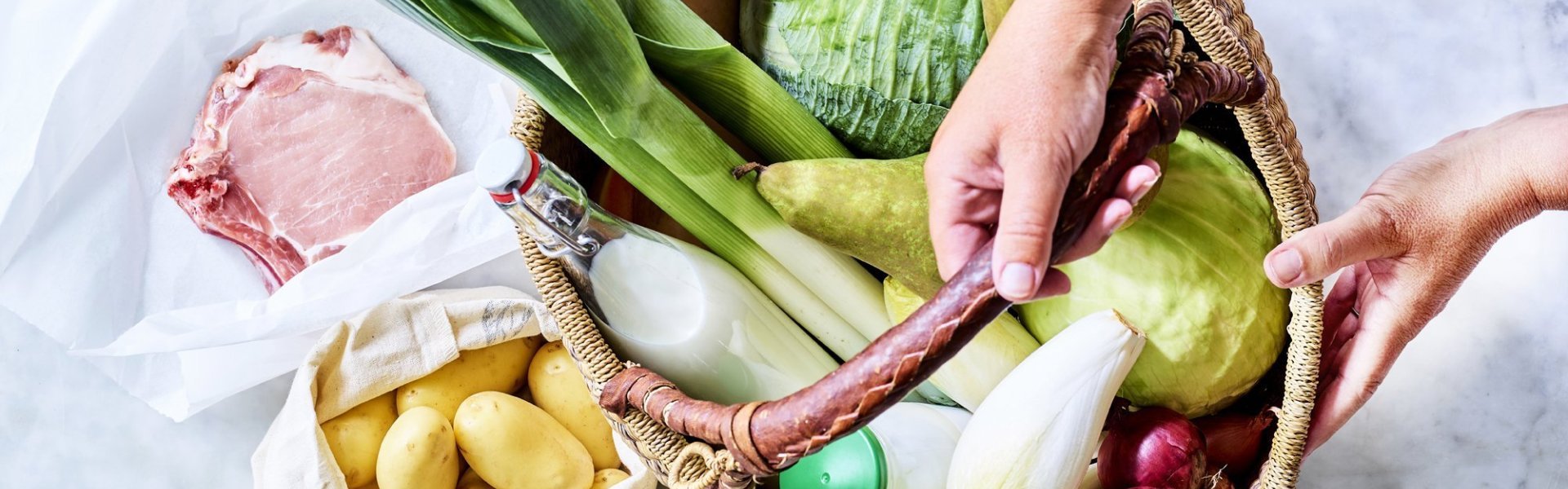 Mand gevuld met producten recht van bij de boer Mand staat op het aanrecht en is gevuld met heerlijke producten van eigen bodem. Denk maar aan prei, witte kool, witloof, yoghurt, uien, krieltjes, varkensvlees... Klaar om meteen aan de slag te gaan.