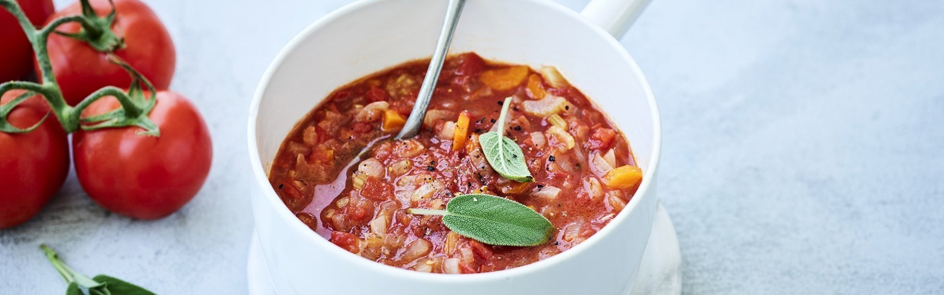 Een klassieke tomaten die is klaargemaakt in een witte kookpot. De tomatensaus zit boordevol verse groenten en tomaten, natuurlijk. Er liggen enkele blaadjes salie op de saus en een verse tros tomaten naast de pot op het marmere aanrecht.