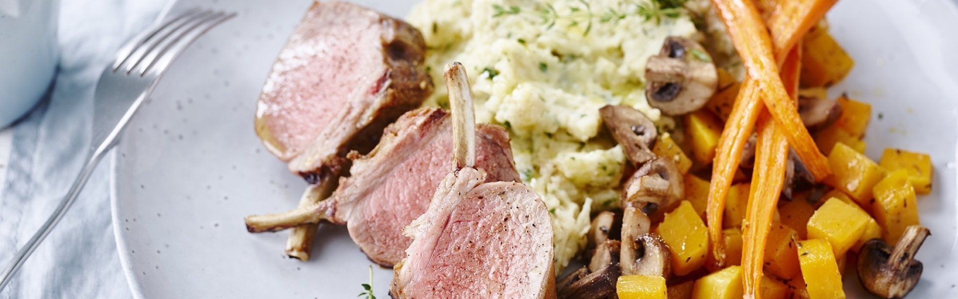 Lamskroon met pastinaakpuree en pompoen in de oven Lamskroontje gesneden in parten, geserveerd met pastinaakpuree, stukjes pompoen, wortel en champignons. Het is een lichtgrijs bord op een lichtblauw linnen tafelkleed. Linksboven zie je verse tijm in bloempot staan.