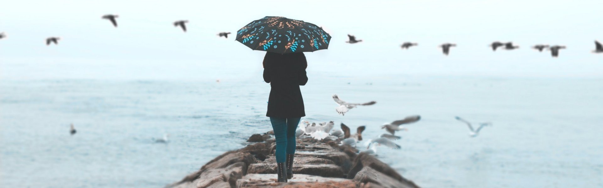 Vrouw met paraplu boven het hoofd loopt op een golfbreker naar de kustlijn terwijl zeemeeuwen vliegen rond haar.