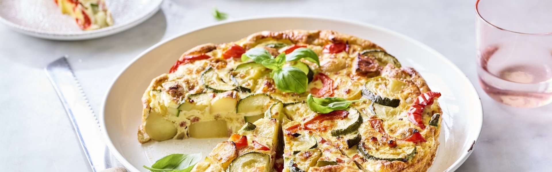 Tortilla met courgette, paprika en ham Een heerlijke aardappeltortilla ligt aangesneden op een groot wit bord. Er werden al twee stukjes uitgesneden en liggen geserveerd op kleine bordjes en afgewerkt met basilicum. Er staan licht gekleurd roze glazen langs het gerecht om de foto nog meer kleur te geven.