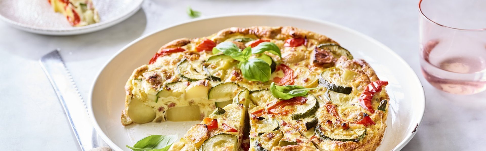 Een heerlijke aardappeltortilla ligt aangesneden op een groot wit bord. Er werden al twee stukjes uitgesneden en liggen geserveerd op kleine bordjes en afgewerkt met basilicum. Er staan licht gekleurd roze glazen langs het gerecht om de foto nog meer kleur te geven.