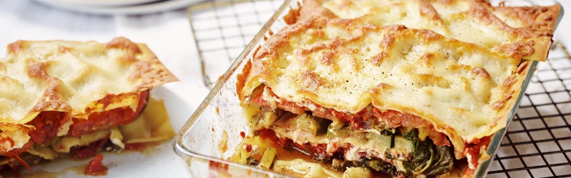 De lasagne staat op tafel in een glazen doorzichtige ovenschotel. Hij is aangesneden zodat je mooi de laagjes ziet van pasta, groenten en saus. De portie die is uitgeschept ligt op een bordje er naast.