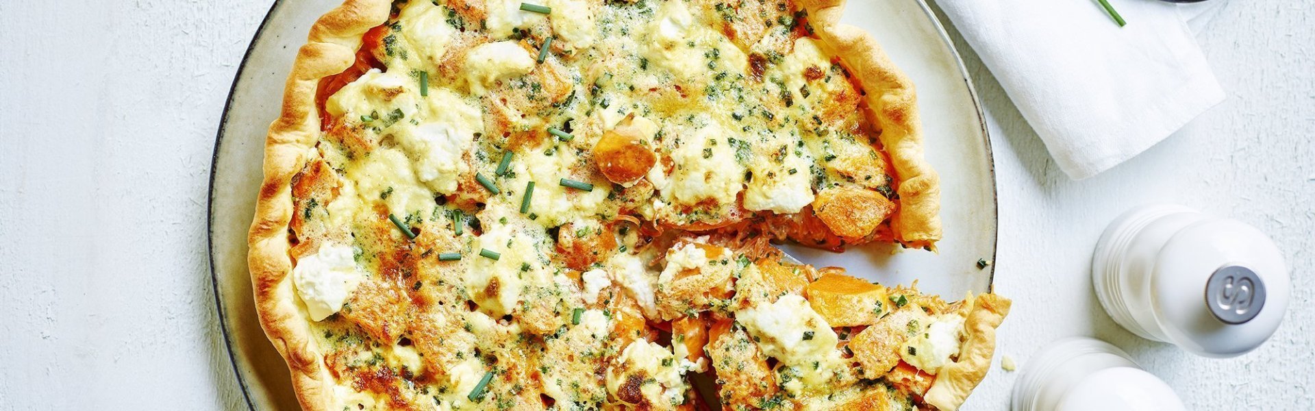 Zoete aardappeltaart met geitenkaas Quiche of groententaart van zoete aardappel met verse geitenkaas. Aangesneden op een wit bord met zwart randje en zilveren taartschep. Links van het bord staan witte molens van peper en zout en rechtsboven liggen enkele bieslooktakjes met keukenschaartje. Dit alles op een witte houten tafel.